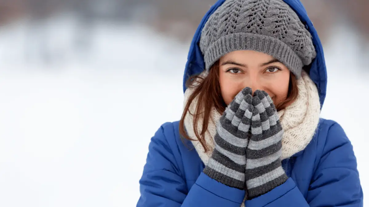Woman in winter coat and knit accessories warming hands outdoors.