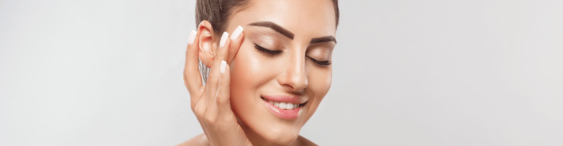 Image of a woman with a serene expression, gently touching her face with her fingers. She has well-groomed eyebrows, long eyelashes, and a healthy complexion, indicative of skincare or beauty treatment. The background is a neutral, light color, emphasizing her flawless skin.