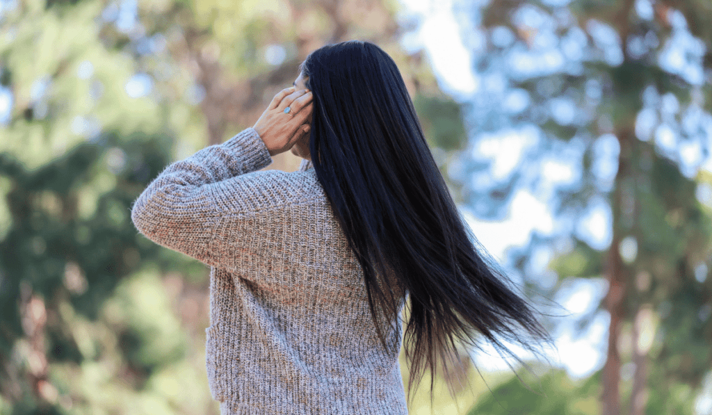 Back view of a woman with long, dark hair standing outdoors, wearing a gray sweater and touching her head.
