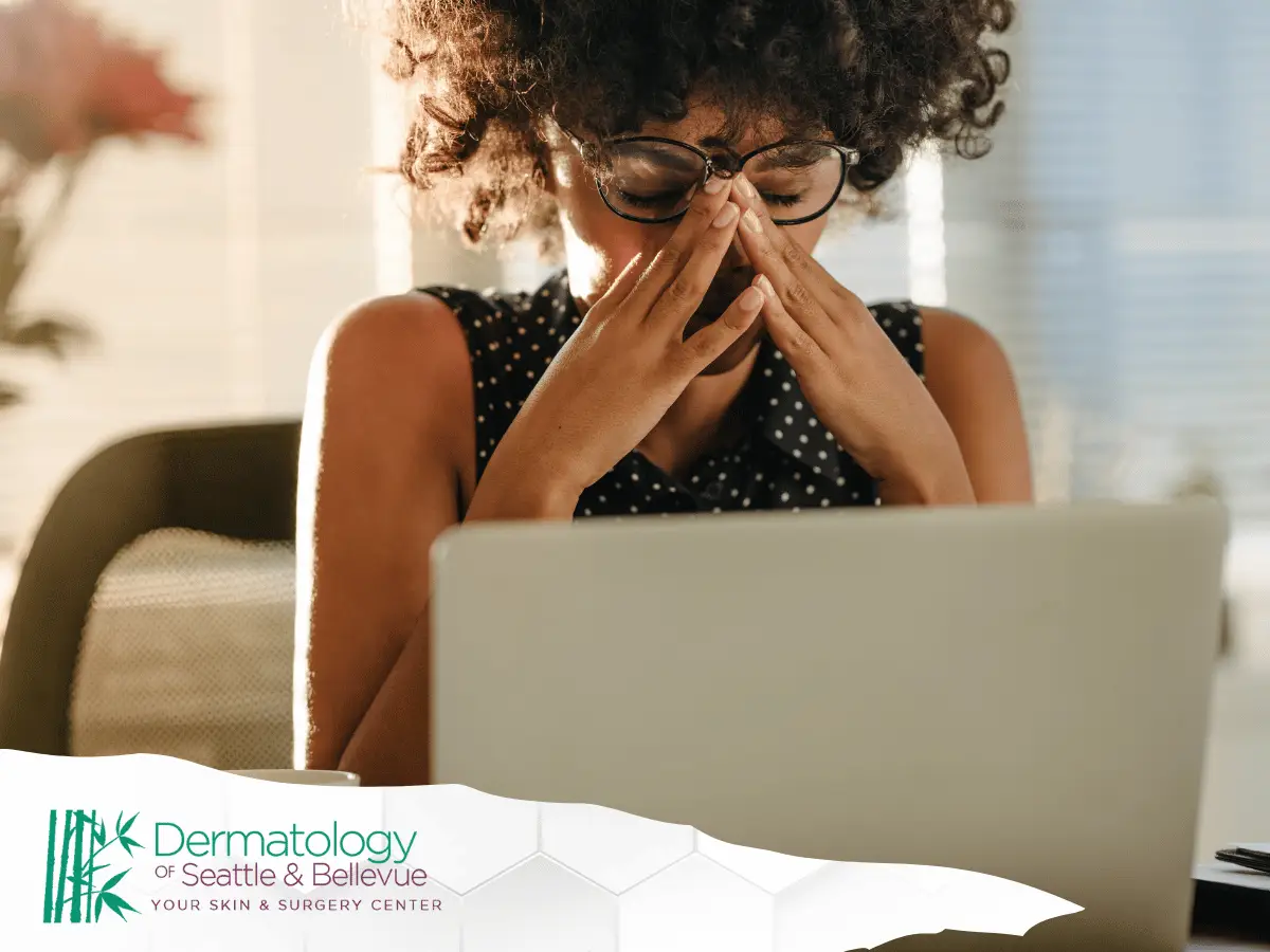A stressed woman wearing glasses sits in front of her laptop, rubbing her temples, with the Dermatology of Seattle & Bellevue logo displayed at the bottom