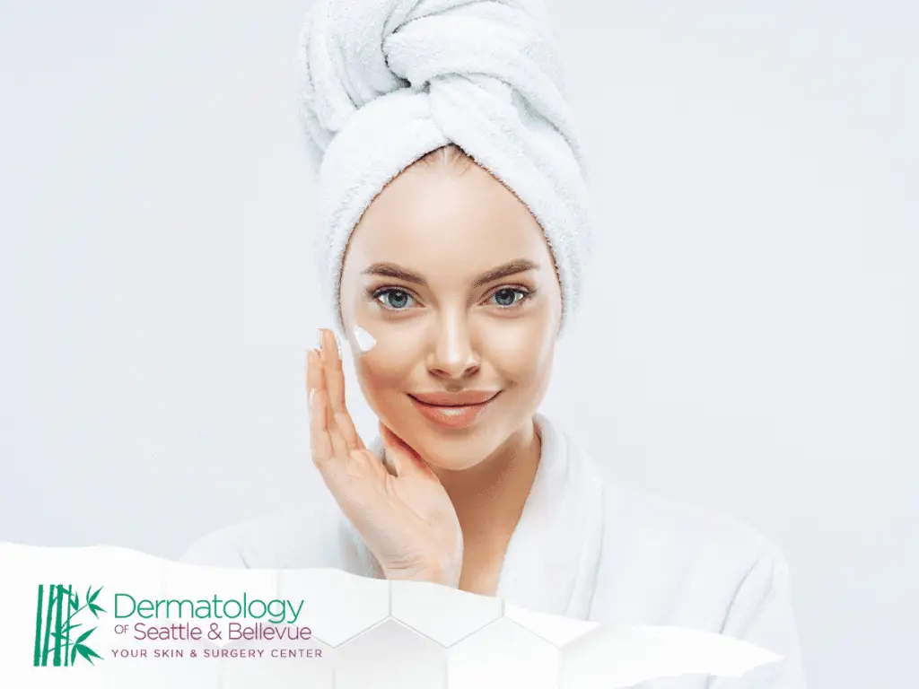 Woman in white towel applying face cream, smiling at camera.