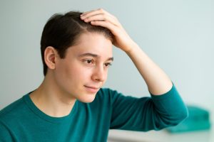 Young man with short brown hair touching his head, looking thoughtful.